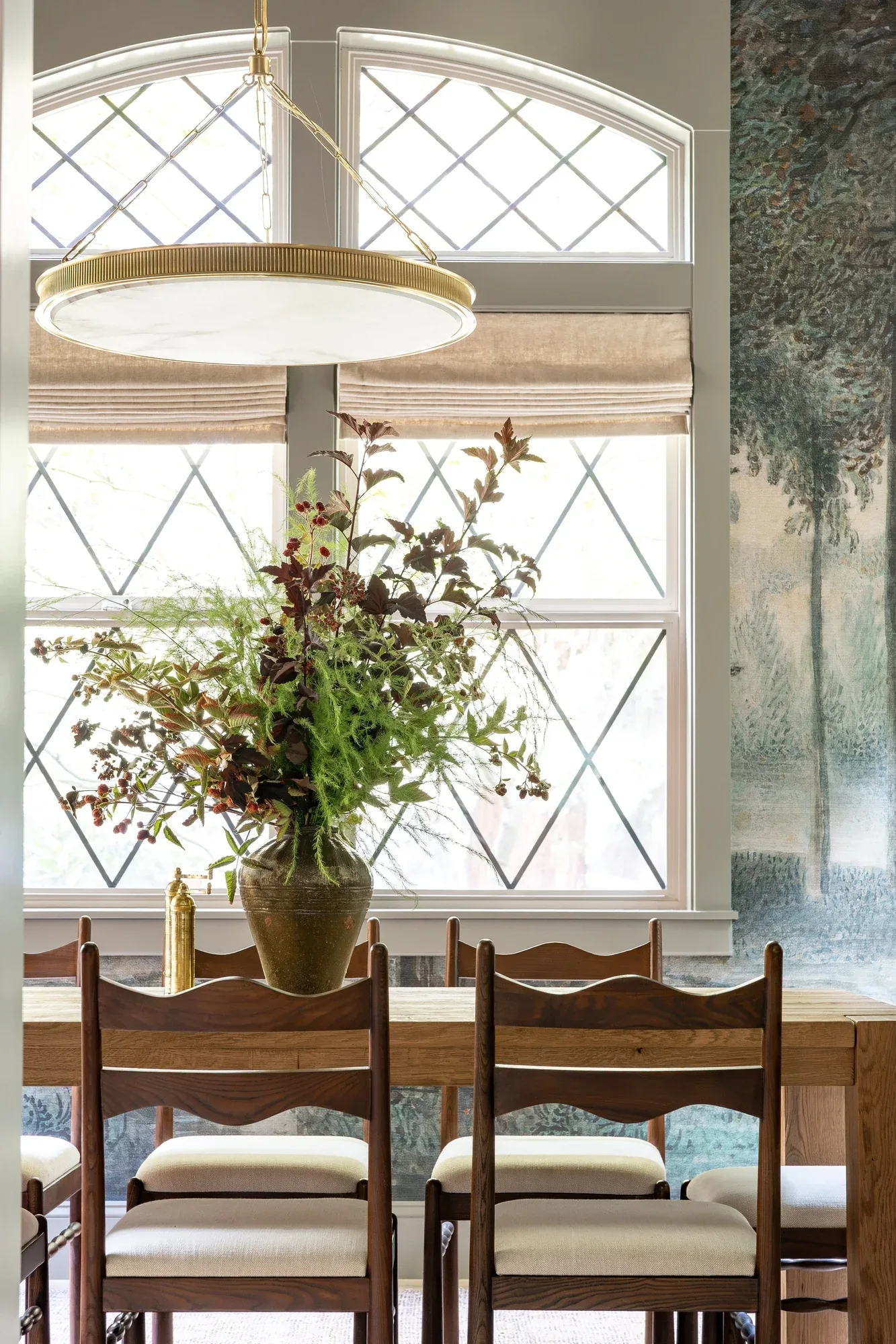A tall arrangement of branches and garden blooms on a dining table beneath leaded arched windows