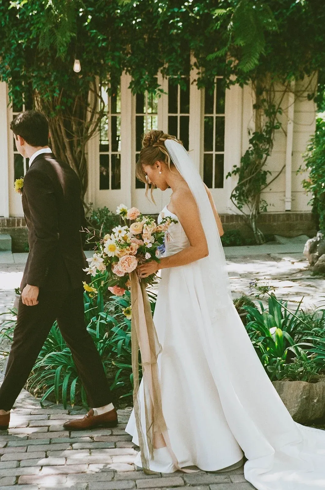 A couple walking through a garden courtyard, the bride carrying a ribboned bouquet